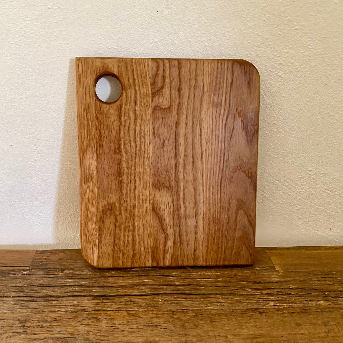 A square oak wooden cutting board with rounded corners and a circular handle cutout in the upper left, standing upright on a wooden surface against a textured off-white wall.