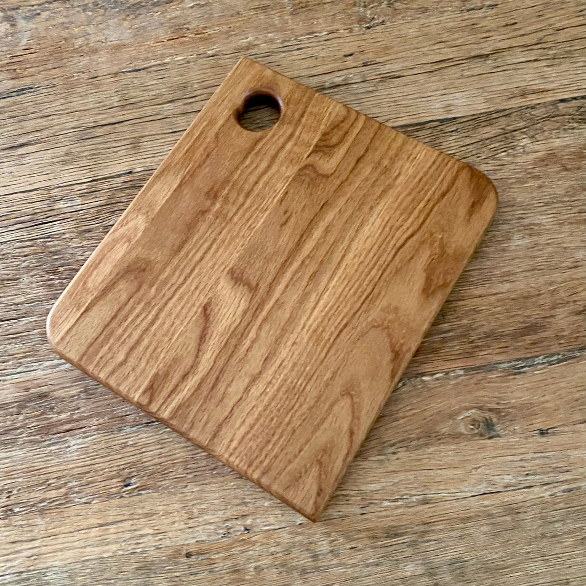 A square white oak cutting board with rounded corners and a circular handle cutout in the top-left corner, shown on a rustic wooden tabletop.