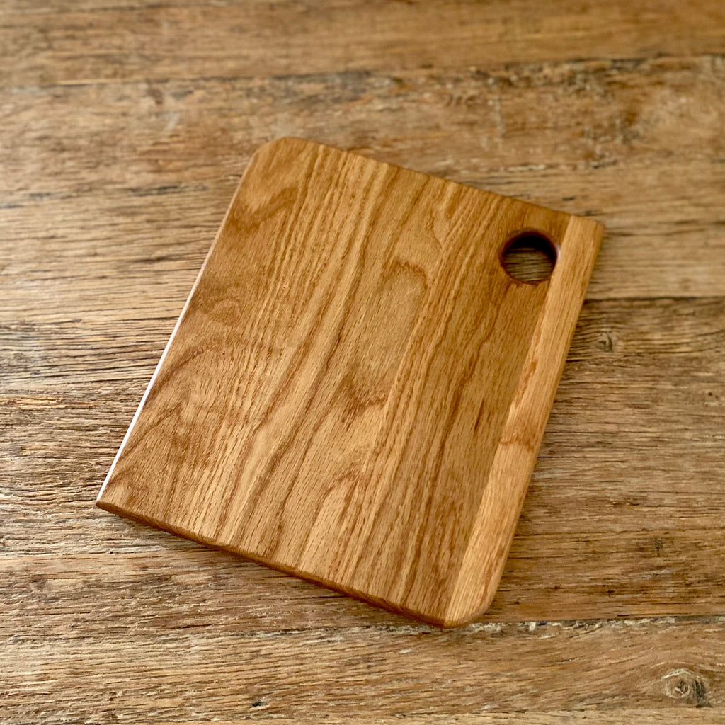 A rectangular wooden white oak cutting board with rounded corners and a circular handle cutout in the top right, shown on a rustic wooden tabletop.
