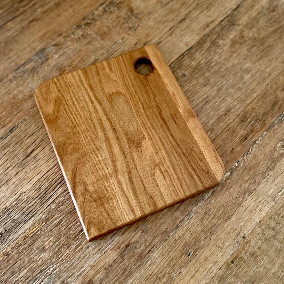 A rectangular white oak cutting board with rounded corners and a small circular hole near one corner, shown on a rustic wooden tabletop.