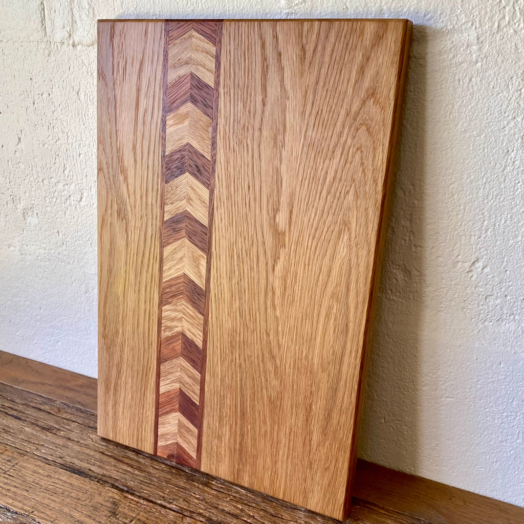 A rectangular white oak and Kwila wooden chopping board with a natural wood grain finish and a central chevron inlay made of alternating light and dark wood tones, leaning against a textured off-white wall on a wooden surface.
