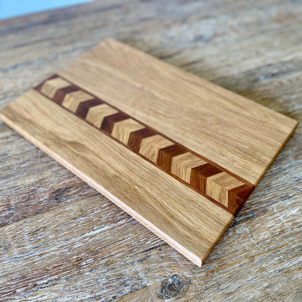 A rectangular wooden chopping board with a natural oak finish and a central chevron inlay made of alternating light and dark wood strips, shown on a rustic wooden tabletop.