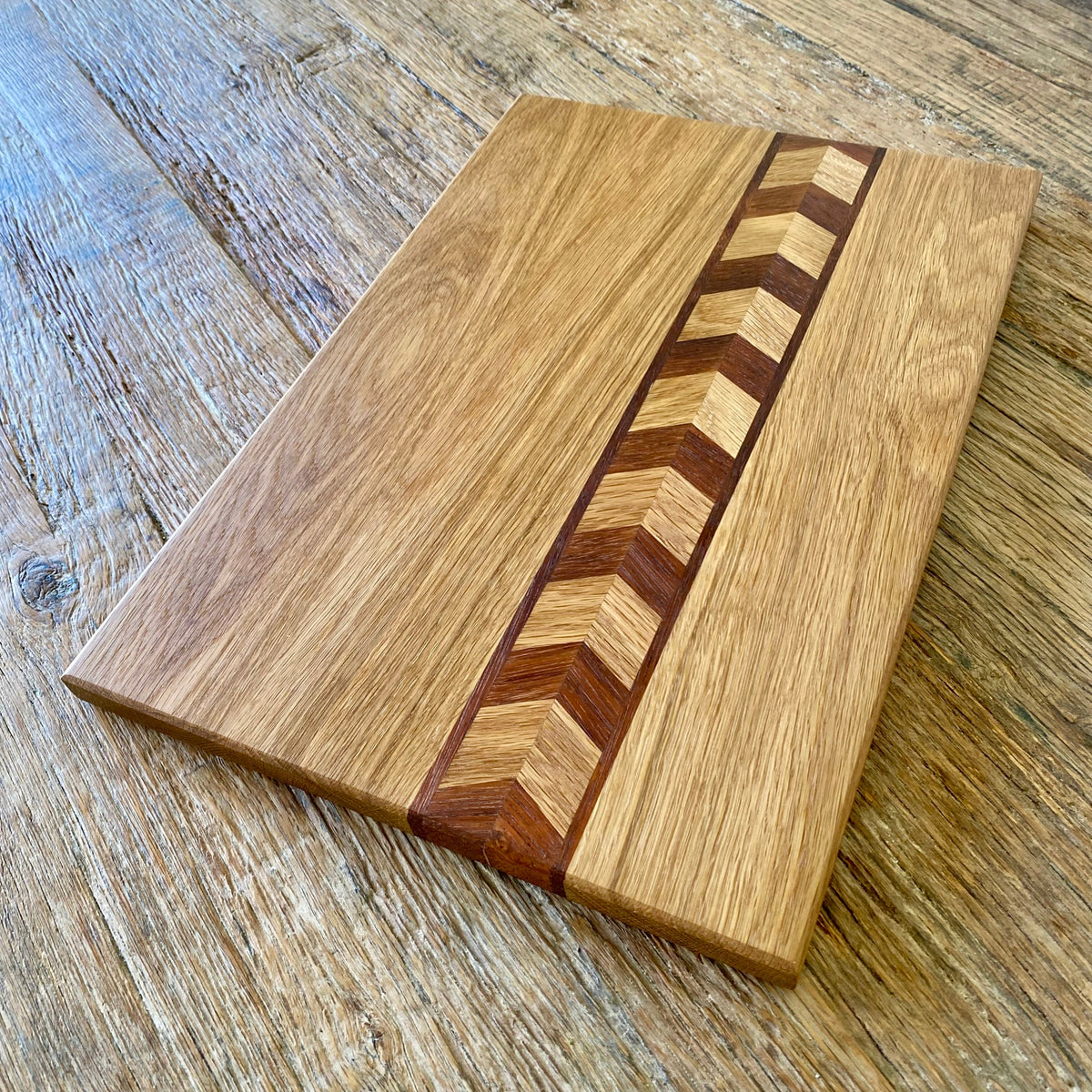 A rectangular wooden cutting board with a natural oak finish and a central chevron inlay of contrasting dark and light wood, shown on a rustic wooden tabletop.