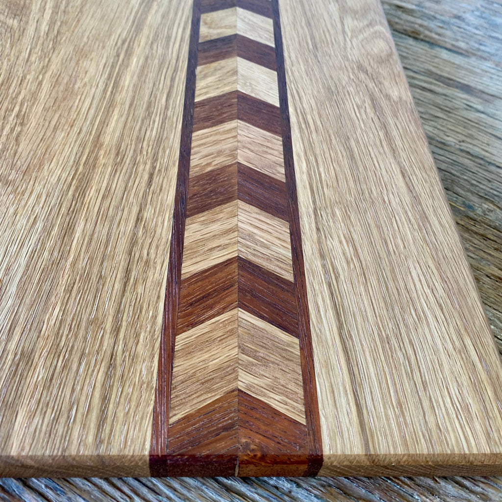 Close-up of a white oak wooden chopping board with a central chevron inlay made of alternating light and dark wood tones, set on a rustic wood surface.