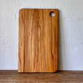 A rectangular wooden Rimu cutting board with rounded corners and a circular hole in the top right, standing upright against a textured off-white wall on a wooden surface.