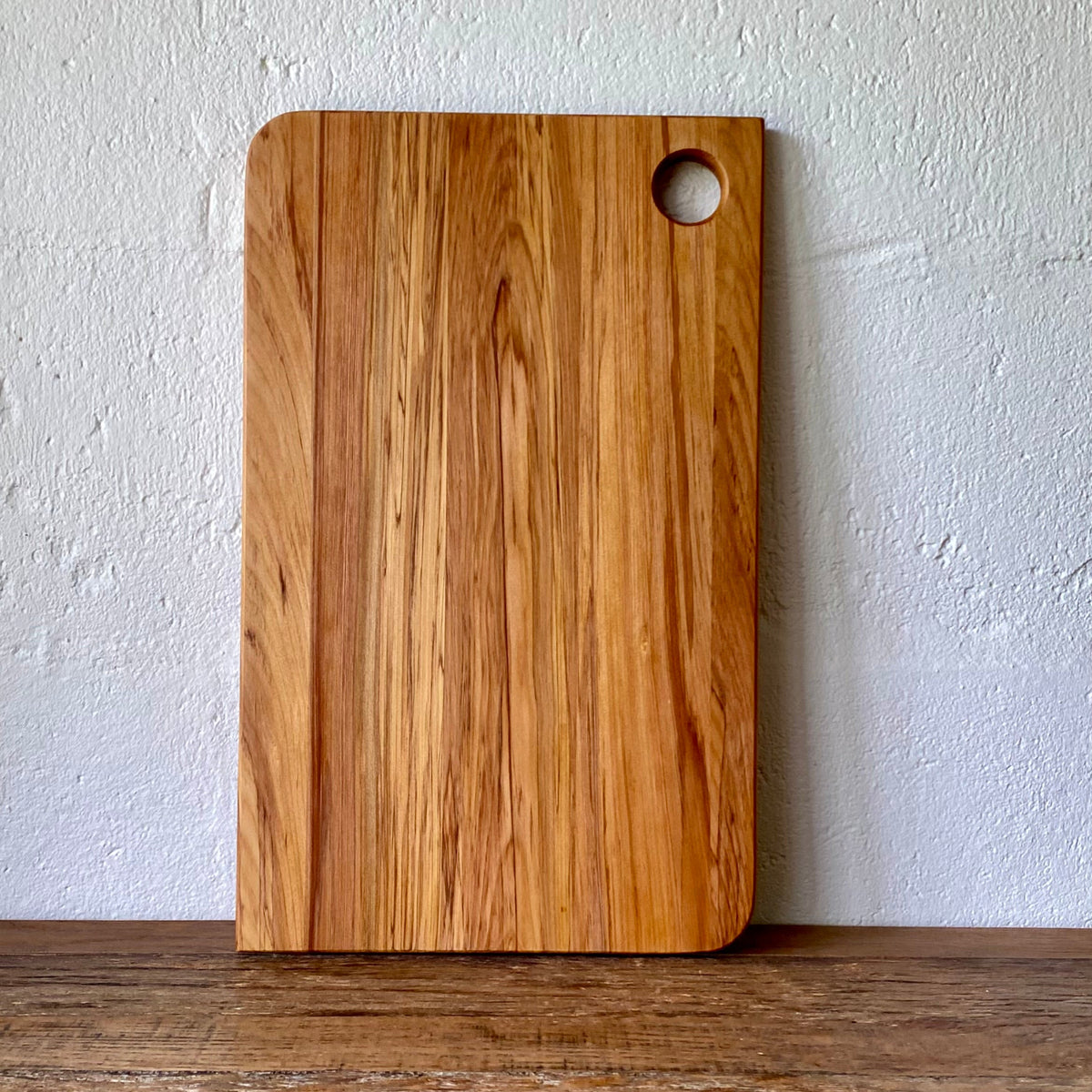 A rectangular wooden Rimu cutting board with rounded corners and a circular hole in the top right, standing upright against a textured off-white wall on a wooden surface.