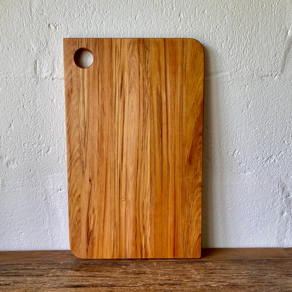 A rectangular wooden Rimu cutting board with rounded corners and a circular hole near the top left, standing upright on a wooden surface against a textured white wall.