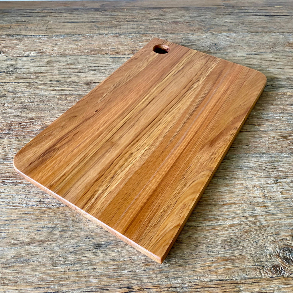 A rectangular wooden Rimu cutting board with rounded corners and a small hole near one end, shown on a rustic wood surface.