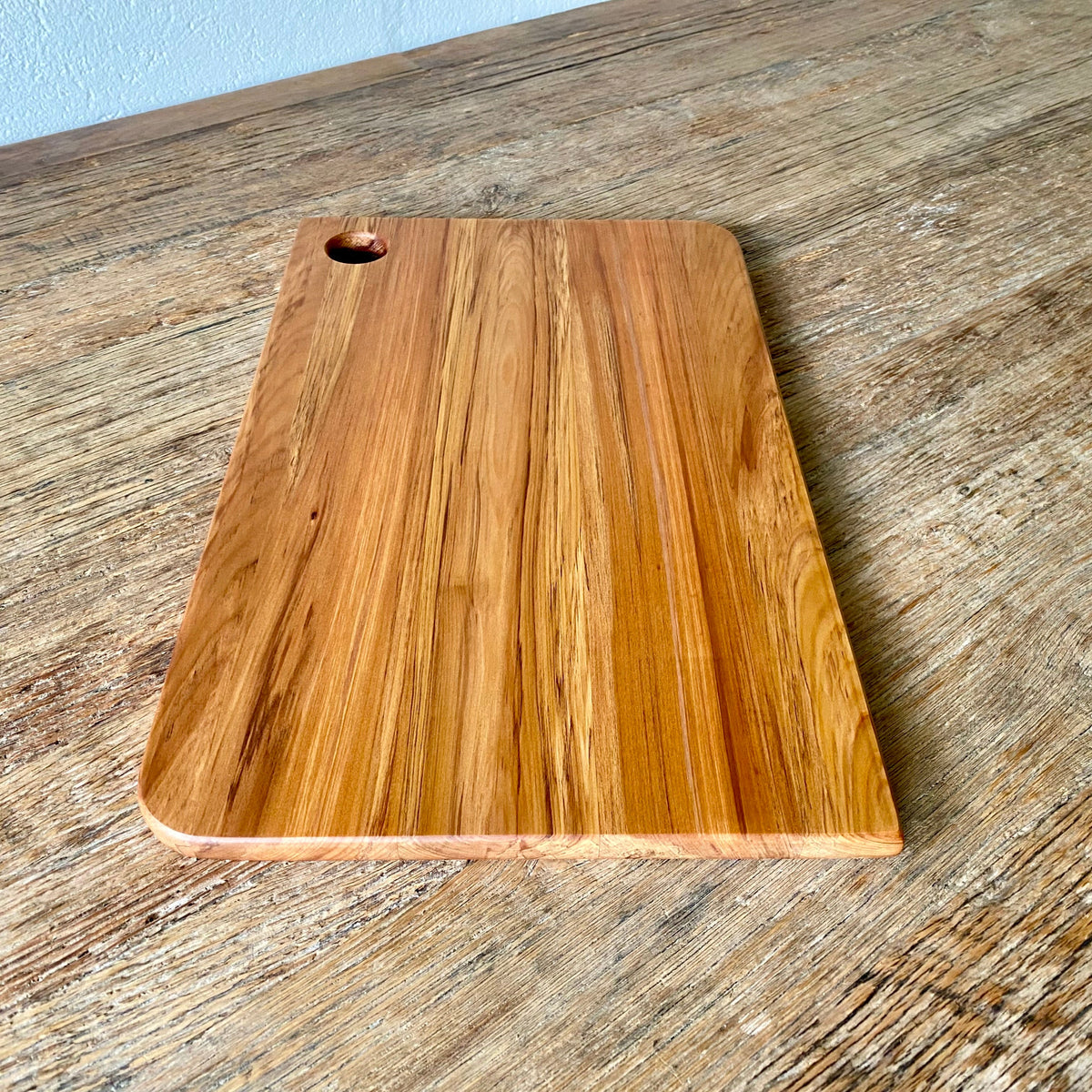 A rectangular wooden Rimu cutting board with rounded corners and a small hanging hole in the top-left corner, placed on a rustic wooden tabletop.