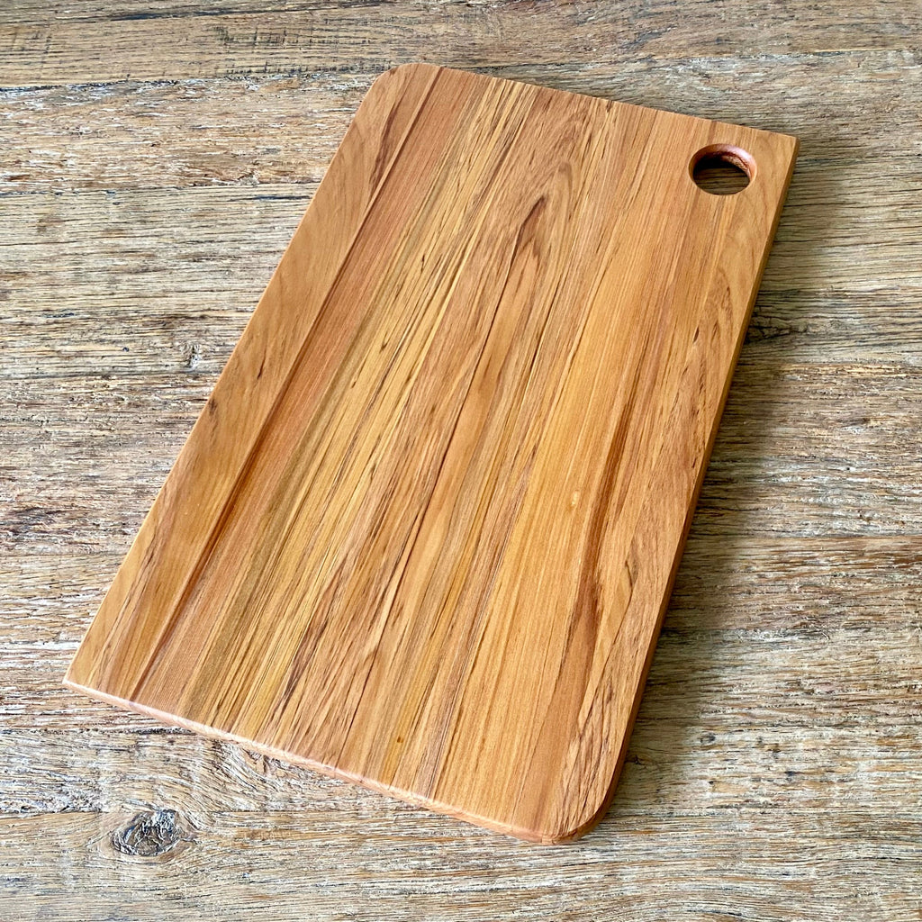 A rectangular wooden Rimu cutting board with rounded corners and a circular handle cutout in the top right, shown on a rustic wooden surface.