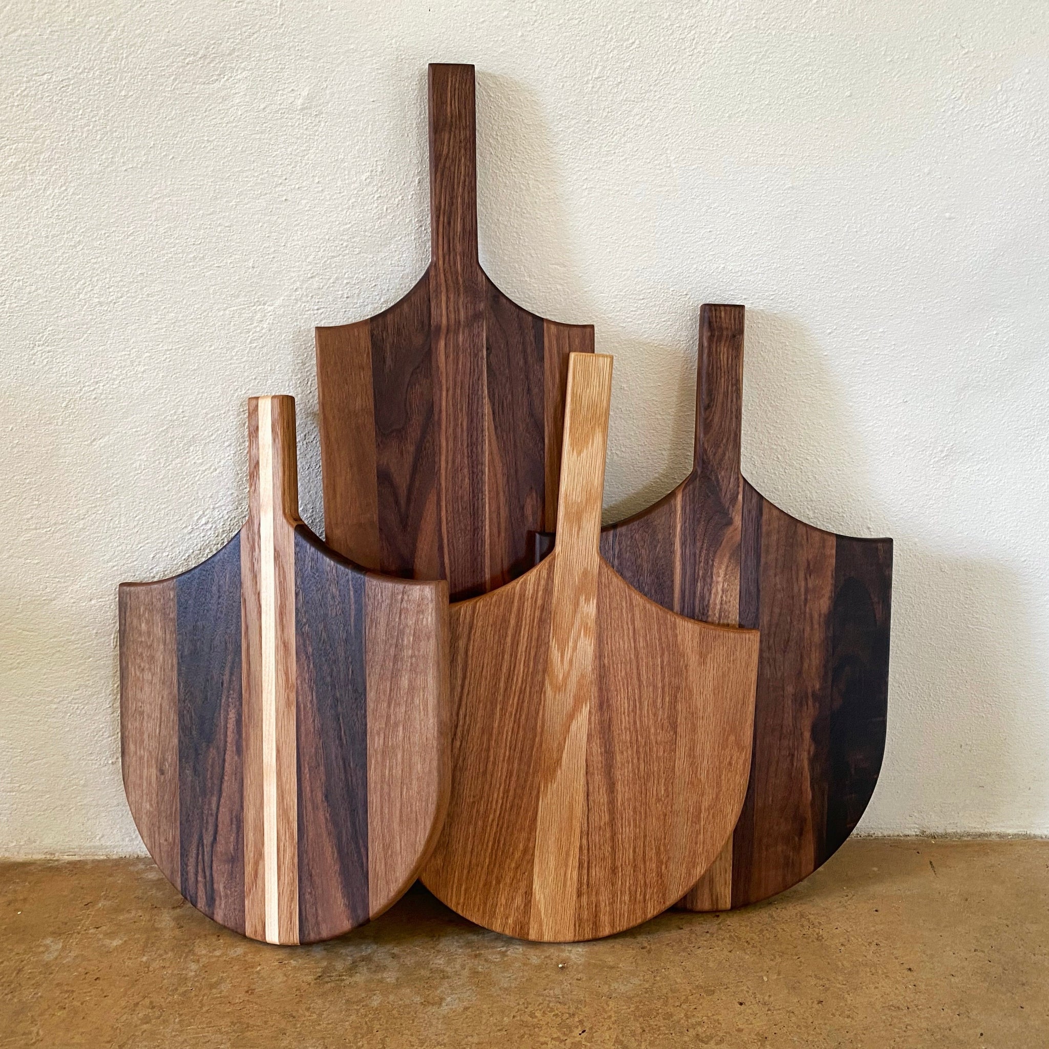 Four wooden paddle-shaped serving boards with different wood grain patterns and colors, arranged against a textured off-white wall.
