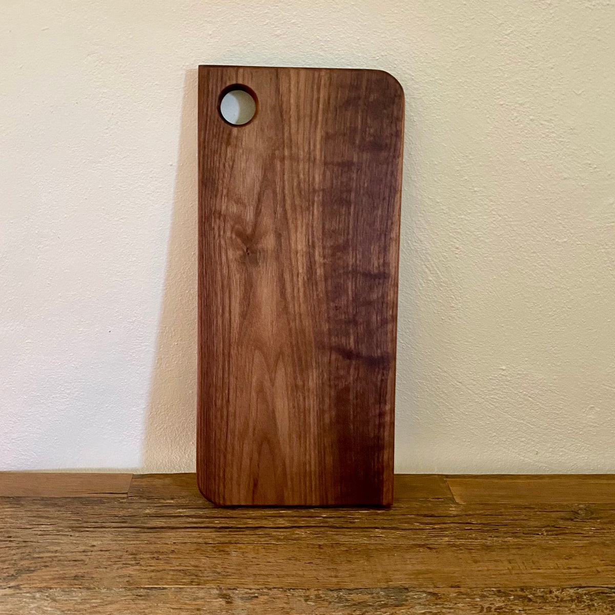 A rectangular wooden cutting board with rounded corners and a circular hole near the top left, standing upright on a wooden surface against a textured off-white wall.