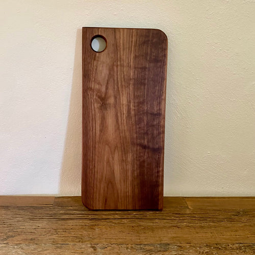 A rectangular wooden cutting board with rounded corners and a circular hole near the top left, standing upright on a wooden surface against a textured off-white wall.