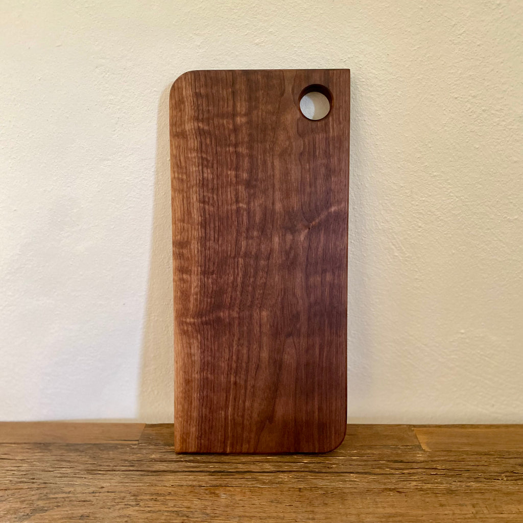 A rectangular dark brown wooden cutting board with rounded corners and a circular hole near the top right, standing on a wooden surface against a light beige wall.