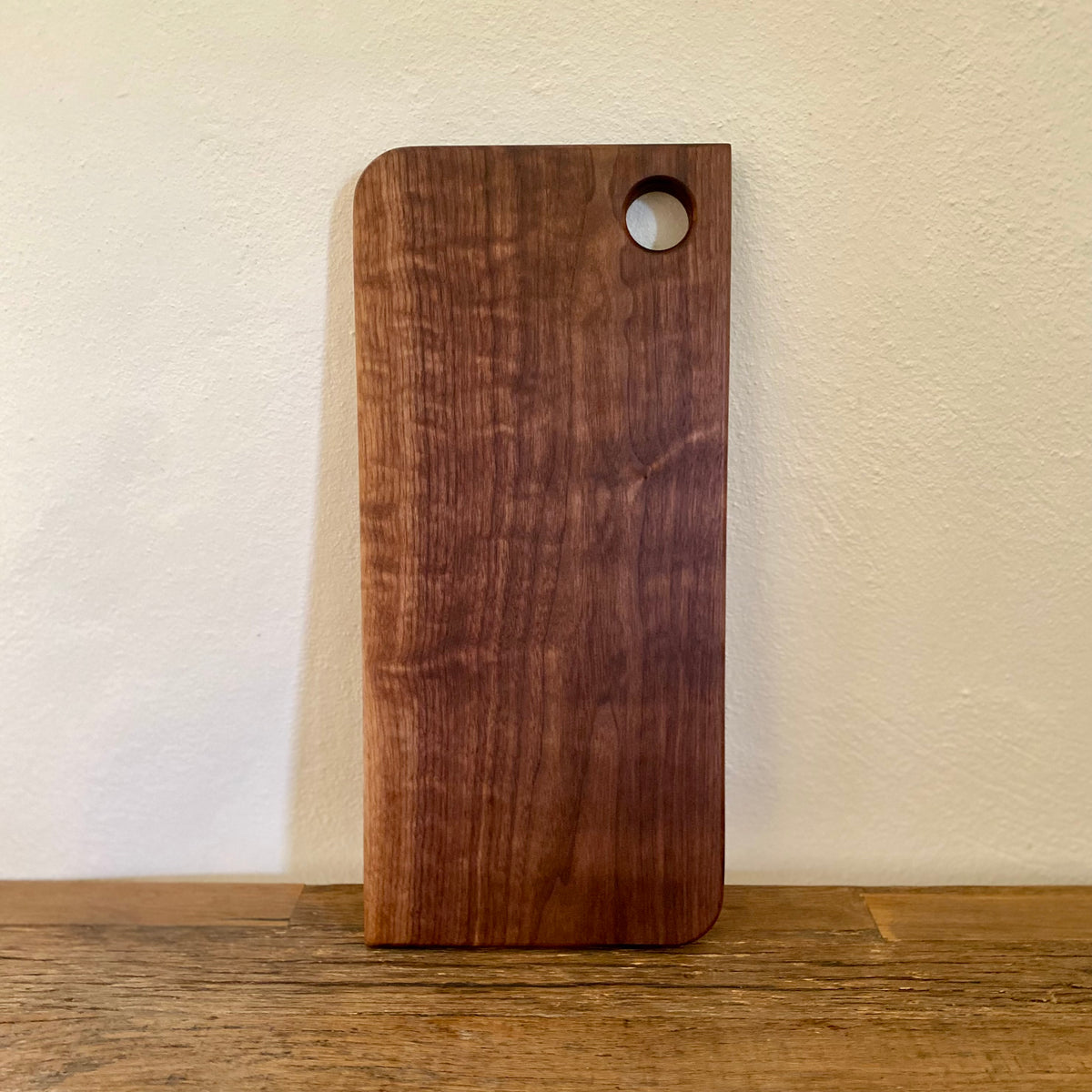 A rectangular dark brown wooden cutting board with rounded corners and a circular hole near the top right, standing on a wooden surface against a light beige wall.