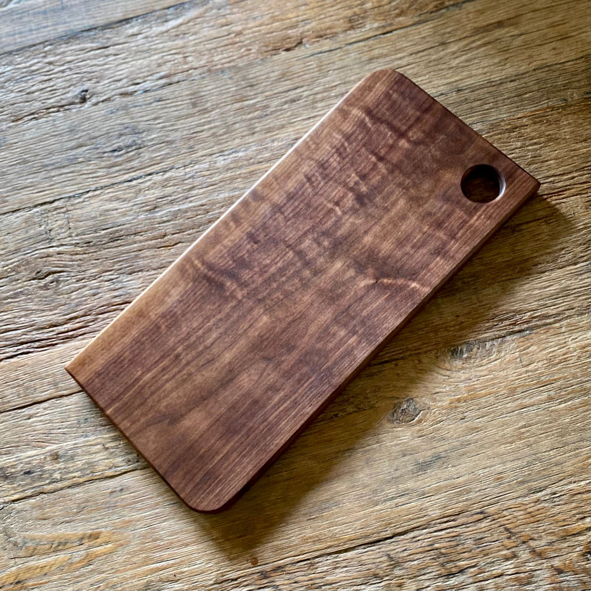 A rectangular wooden cutting board with rounded corners and a small circular hole in the top right corner, resting on a rustic wooden tabletop.