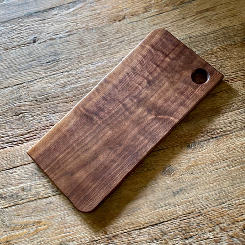 A rectangular wooden cutting board with rounded corners and a small circular hole in the top right corner, resting on a rustic wooden tabletop.