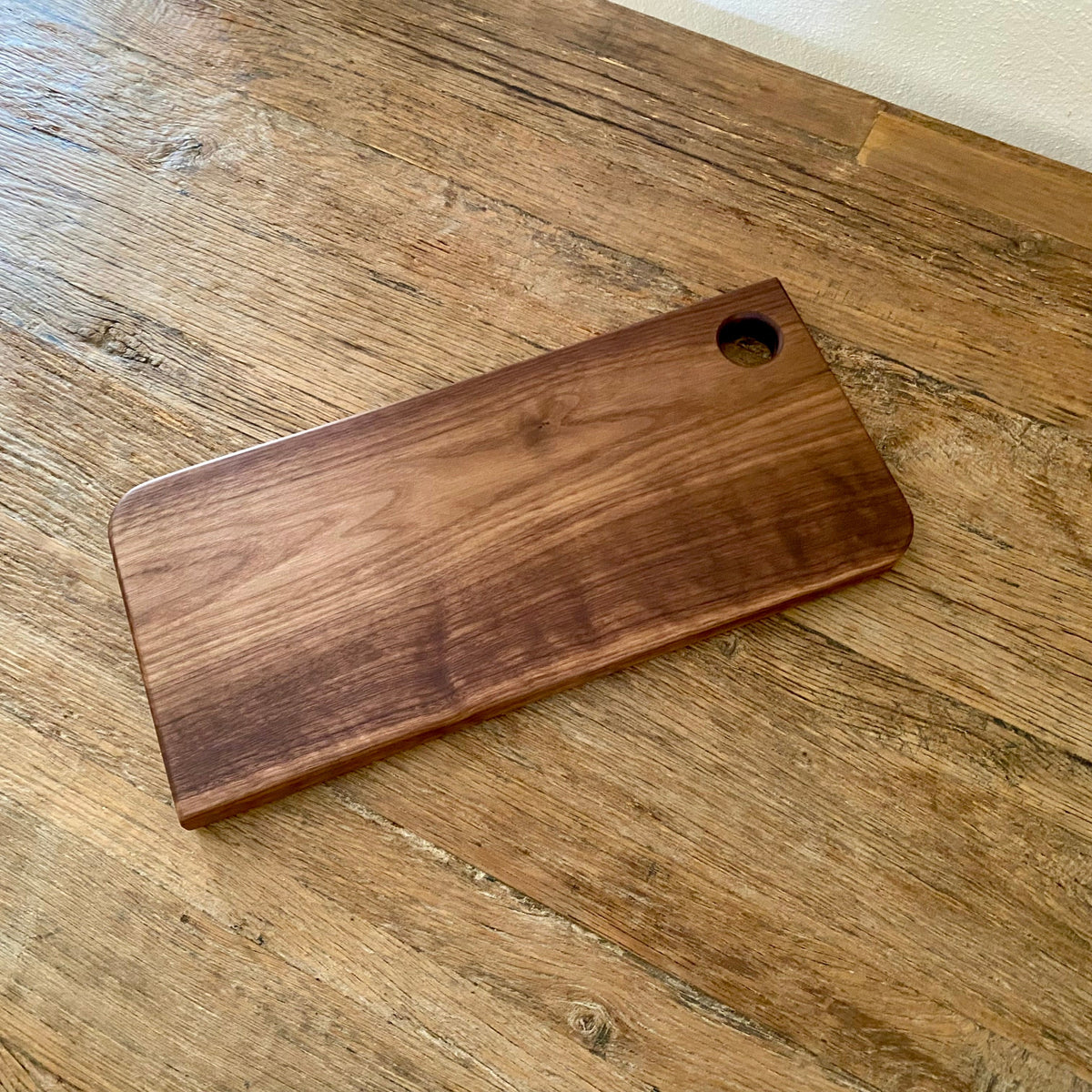 A rectangular wooden cutting board with rounded corners and a circular hole in the top-right corner, resting on a rustic wooden tabletop.