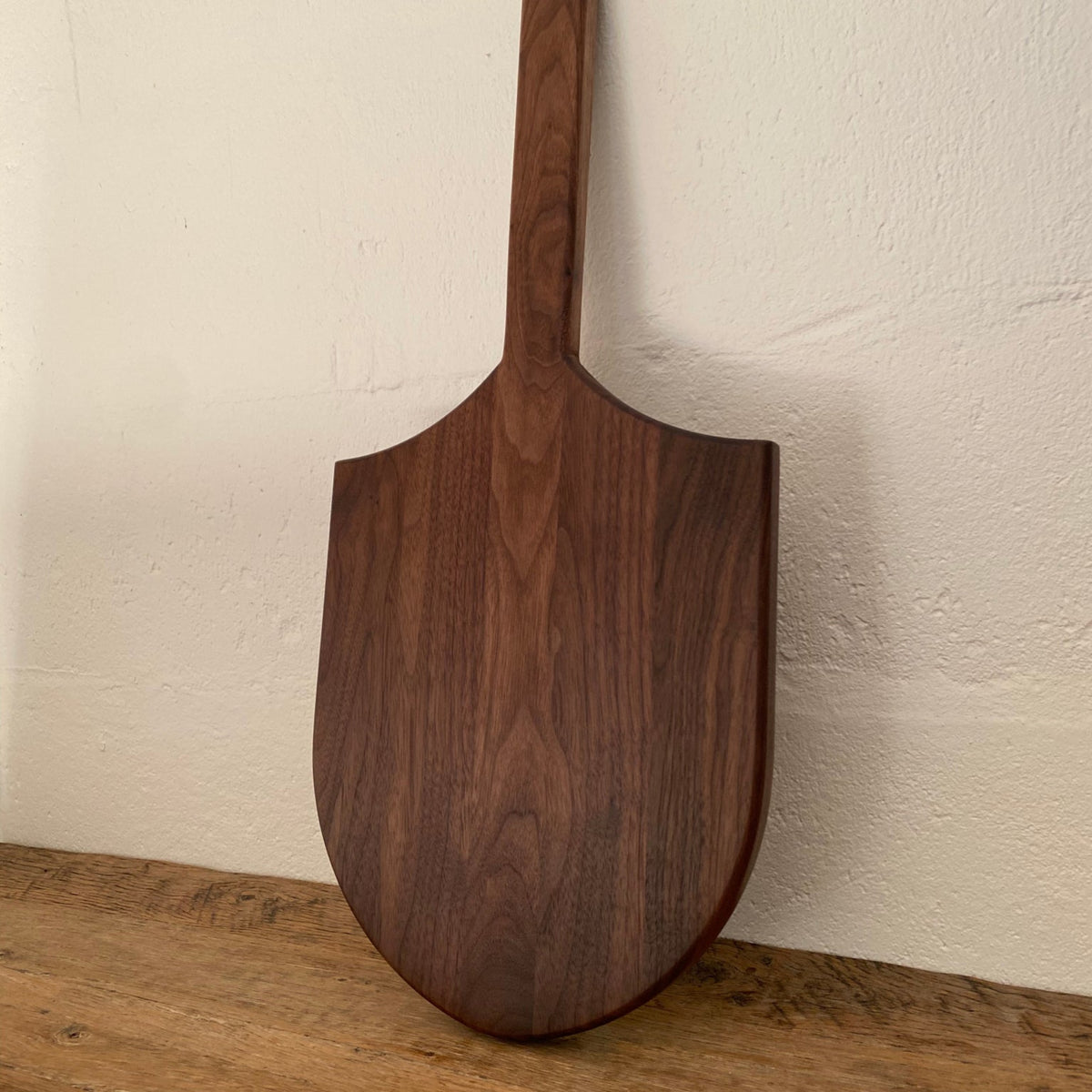Long Tail Ray Board in American walnut – front view showing shield-shaped board and long handle, leaning against white wall