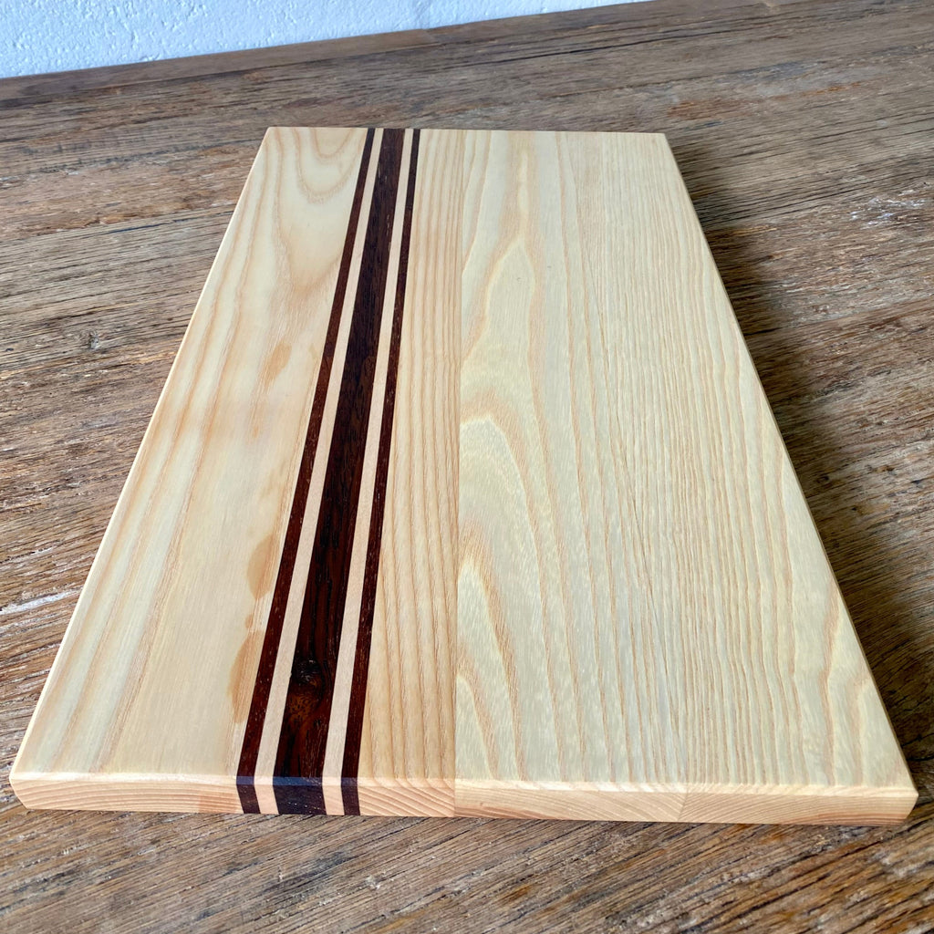 Ash Long platter Board with Kwila and Maple stripes running the length just off centre. showing the end of the board laying on a rustic table