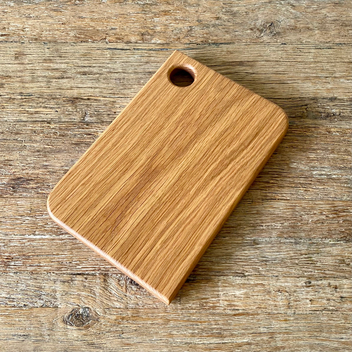 A rectangular white oak wooden cutting board with rounded corners and a small circular hole near one corner, shown on a rustic wooden surface.