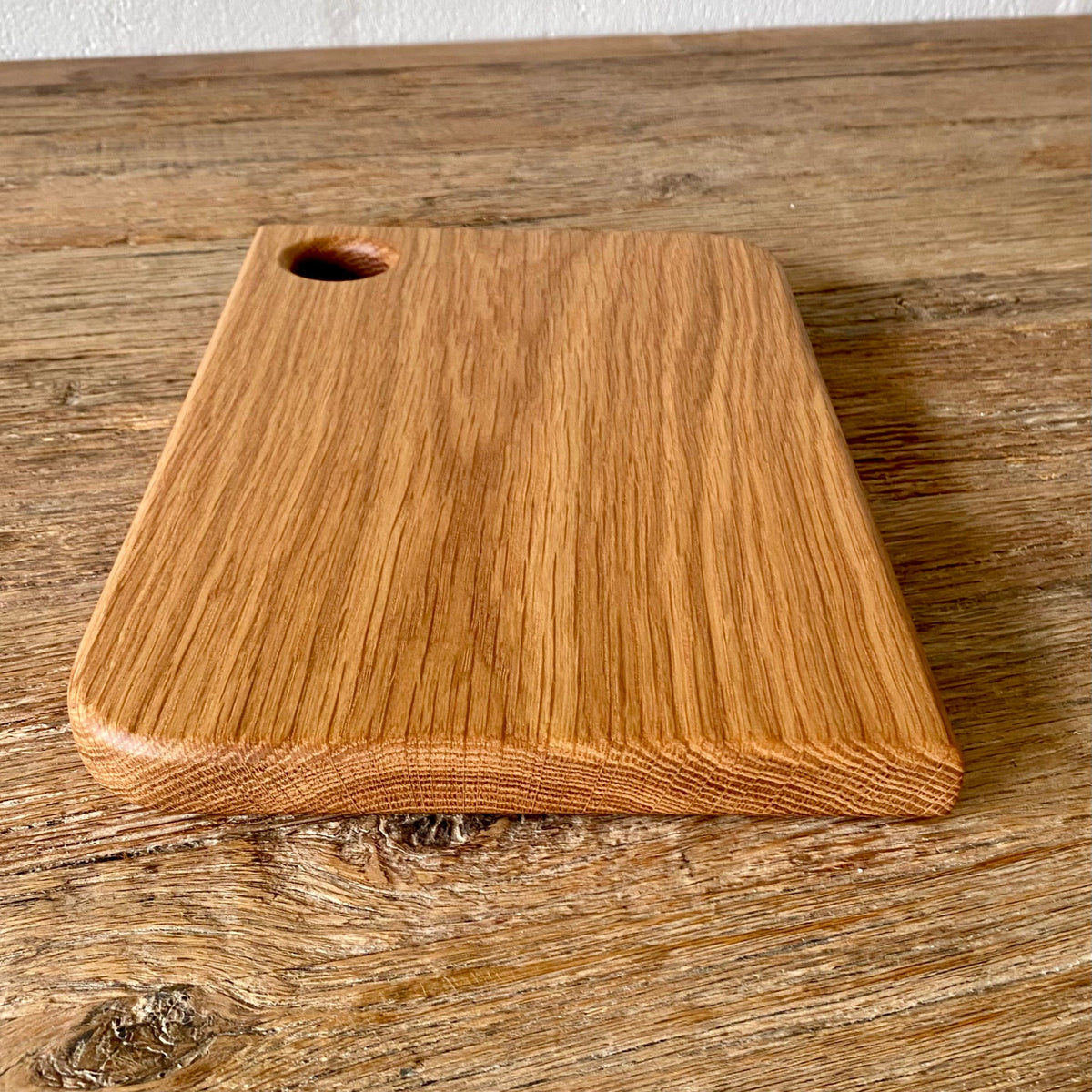 A rectangular white oak wooden cutting board with rounded corners and a small circular hole in the top-left corner, resting on a rustic wooden tabletop against a white wall.
