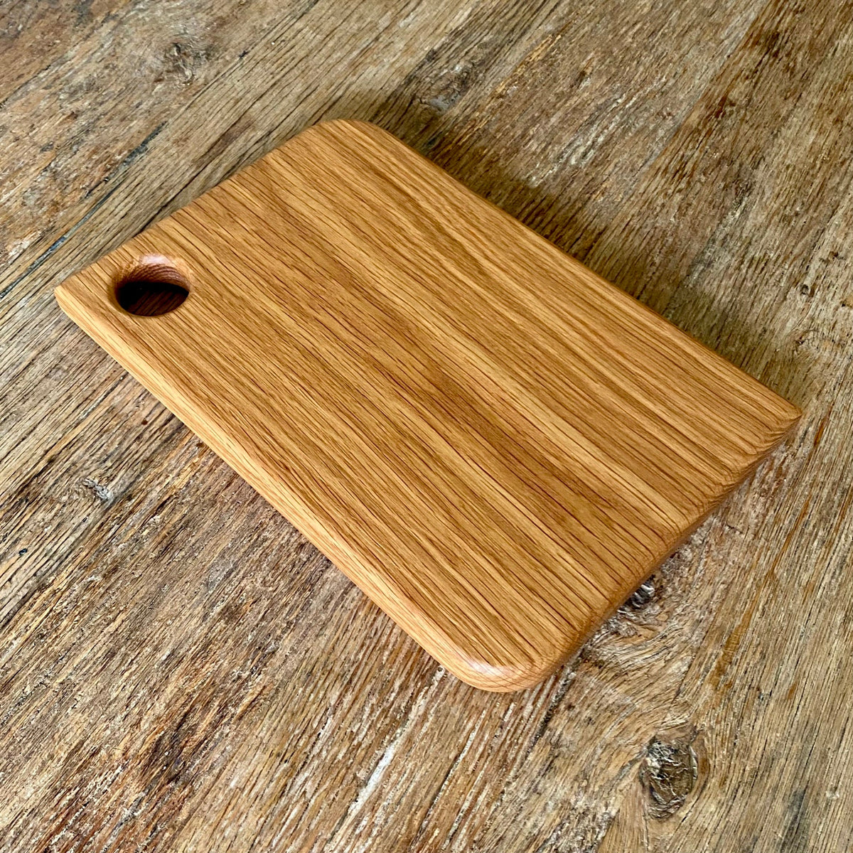 A rectangular white oak wooden cutting board with rounded corners and a small hole in one corner, shown on a rustic wooden tabletop.