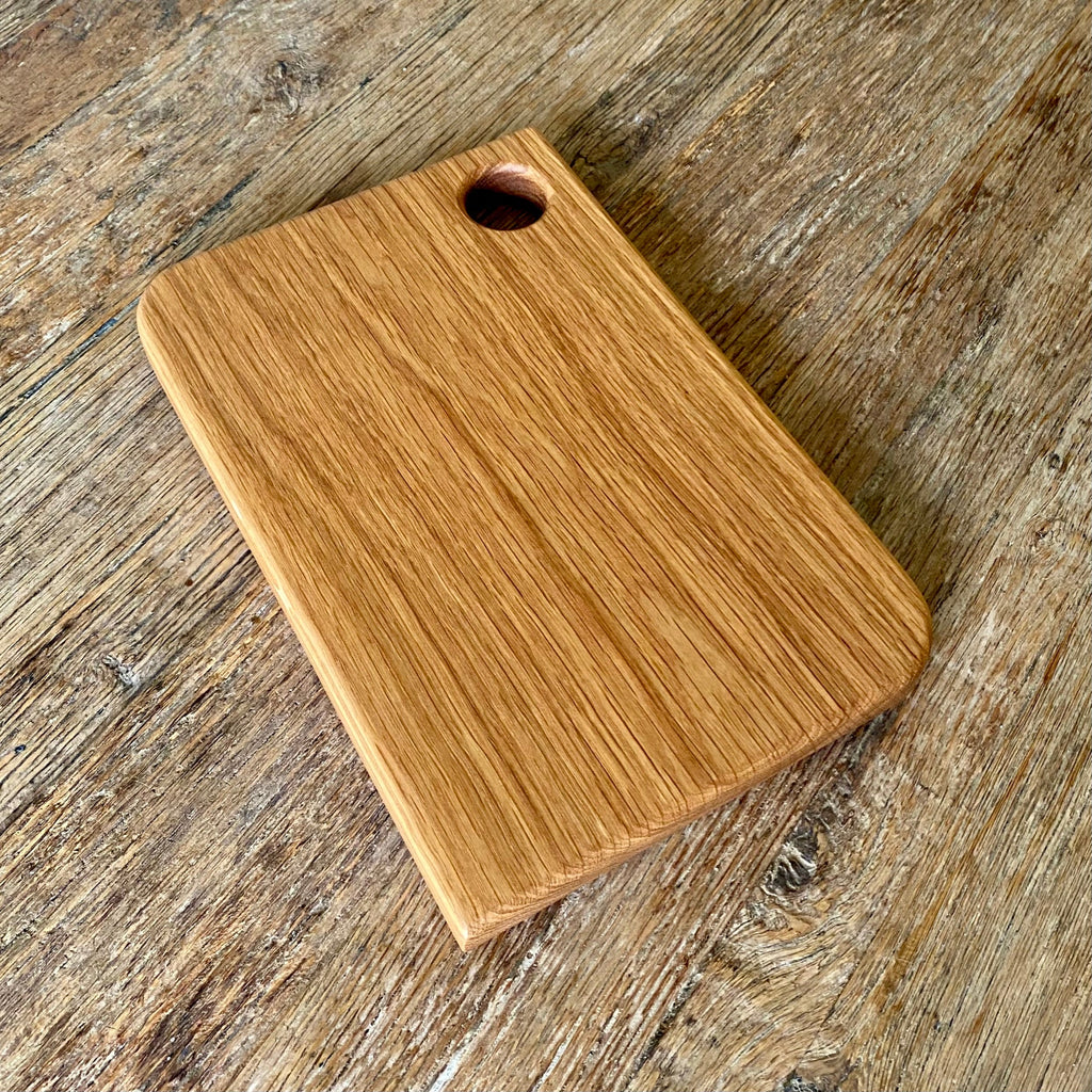 A rectangular white oak  wooden cutting board with rounded corners and a small circular hole near one corner, shown on a rustic wooden surface.