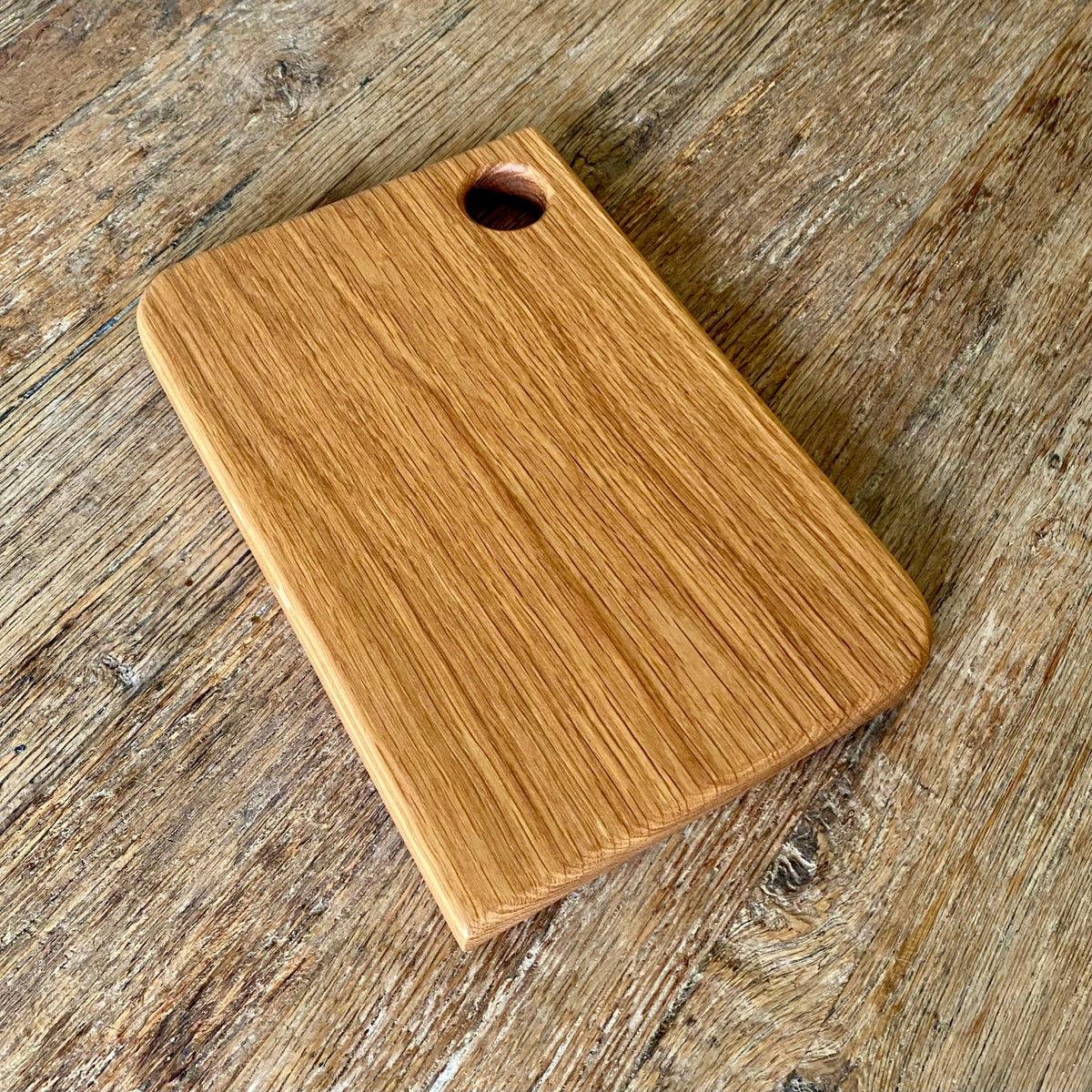 A rectangular white oak  wooden cutting board with rounded corners and a small circular hole near one corner, shown on a rustic wooden surface.