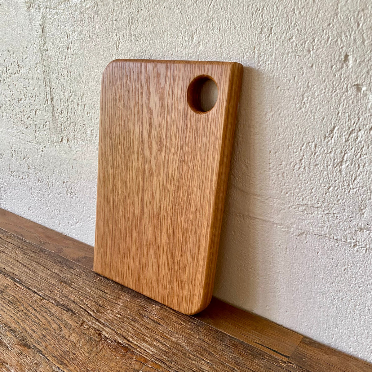A rectangular white oak wooden cutting board with rounded corners and a circular hole near the top right, leaning against a textured off-white wall on a wooden surface.