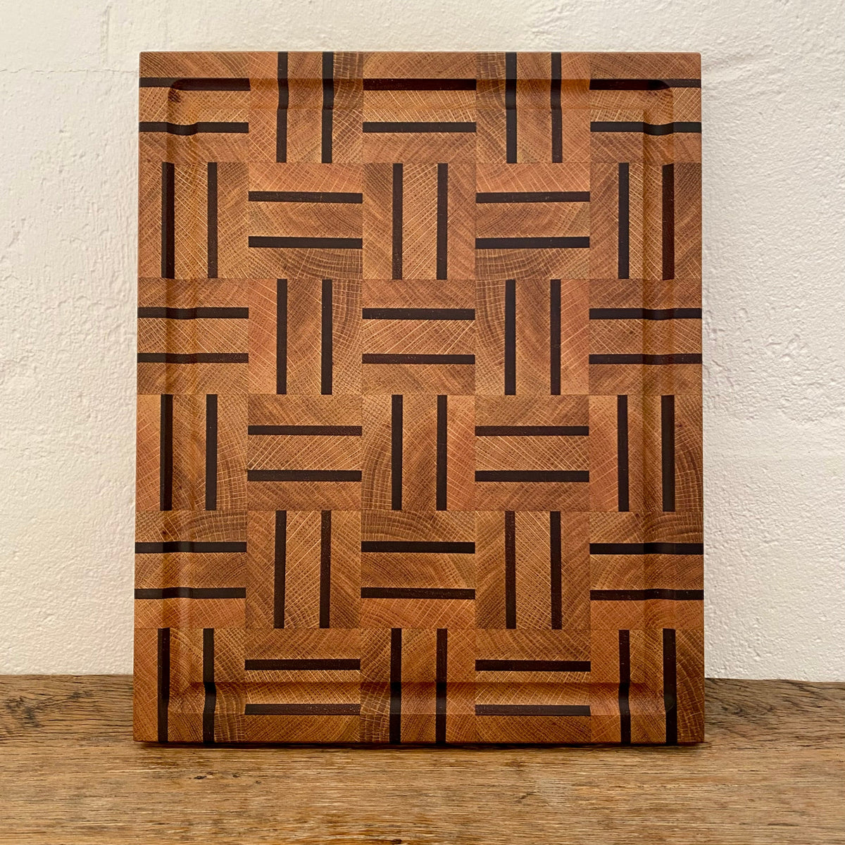 A rectangular white oak kwila wooden cutting board with a geometric inlay pattern of dark brown and medium brown wood blocks, standing on a wooden surface against a textured off-white wall.