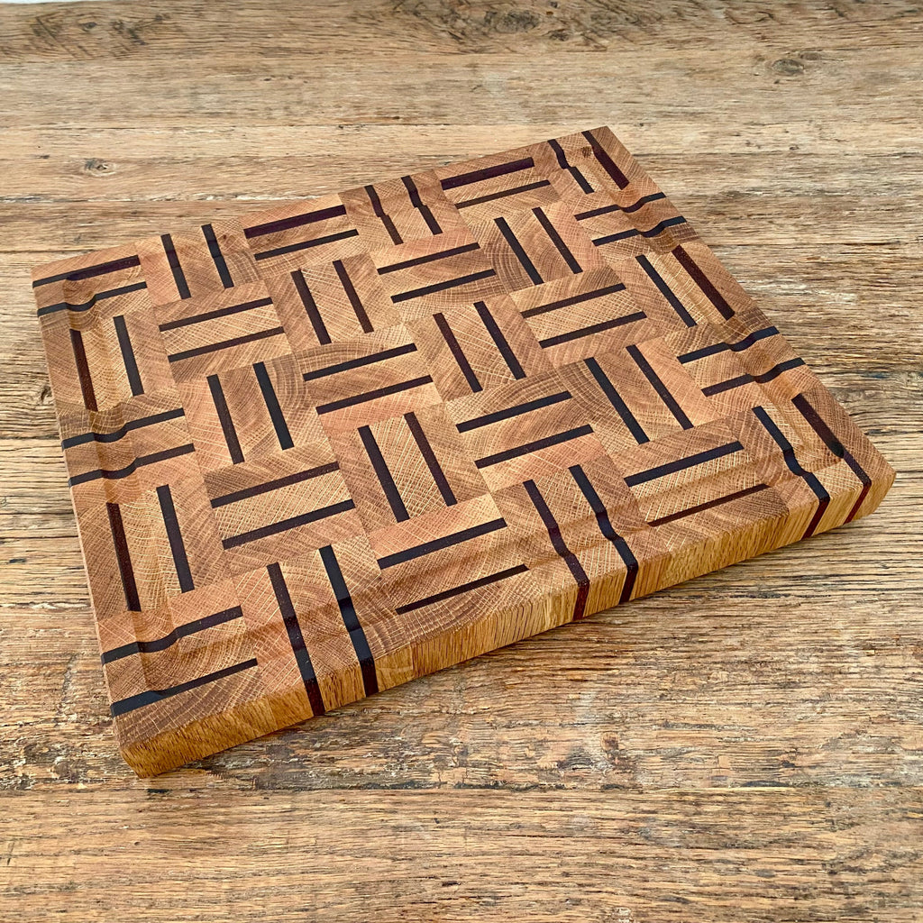 A rectangular white oak and kwila wooden cutting board with a juice groove and geometric inlay pattern of dark brown and medium brown wood blocks, on a rustic wooden table
