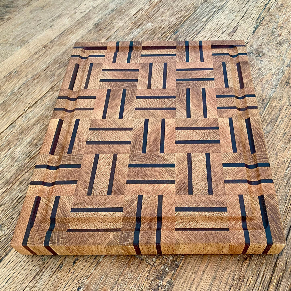 A rectangular white oak and kwila wooden cutting board with a juice groove and geometric inlay pattern of dark brown and medium brown wood blocks, top down photo on a rustic wooden table