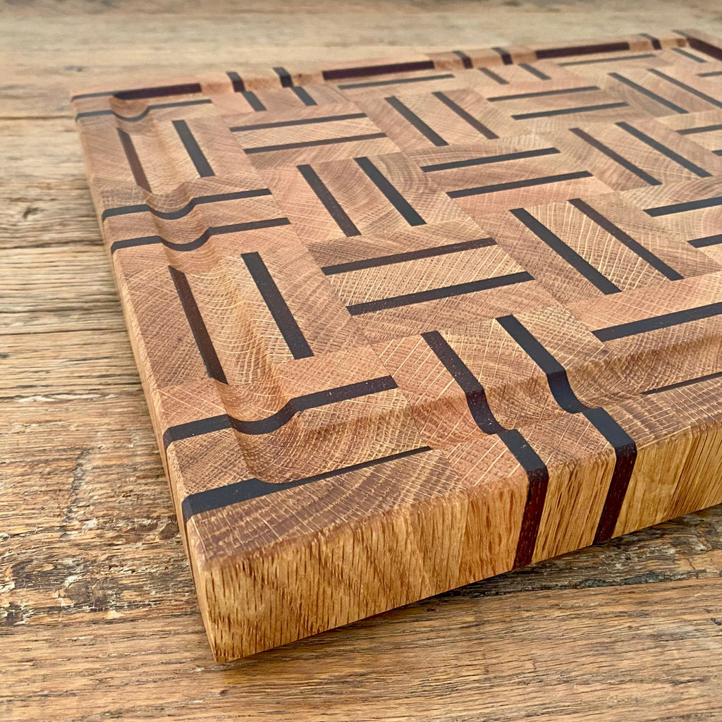 A rectangular white oak and kwila wooden cutting board with a juice groove and geometric inlay pattern of dark brown and medium brown wood blocks, on a rustic wooden table