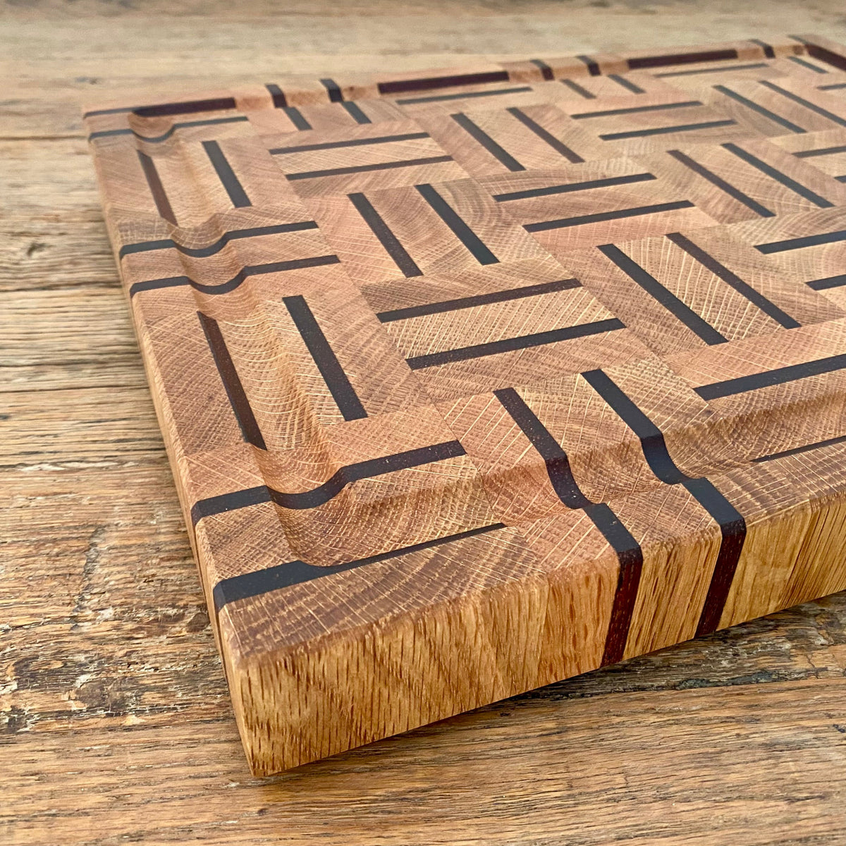 A rectangular white oak and kwila wooden cutting board with a juice groove and geometric inlay pattern of dark brown and medium brown wood blocks, on a rustic wooden table