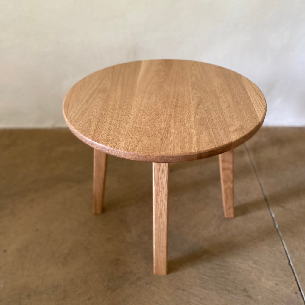 A round wooden side table with a natural oak finish, featuring four tapered legs, set against a neutral concrete floor and textured wall.