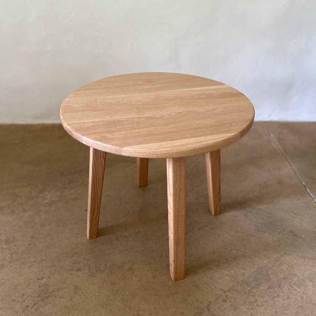 A round wooden side table with a light natural oak finish, featuring four tapered legs, set against a neutral concrete floor and textured wall.