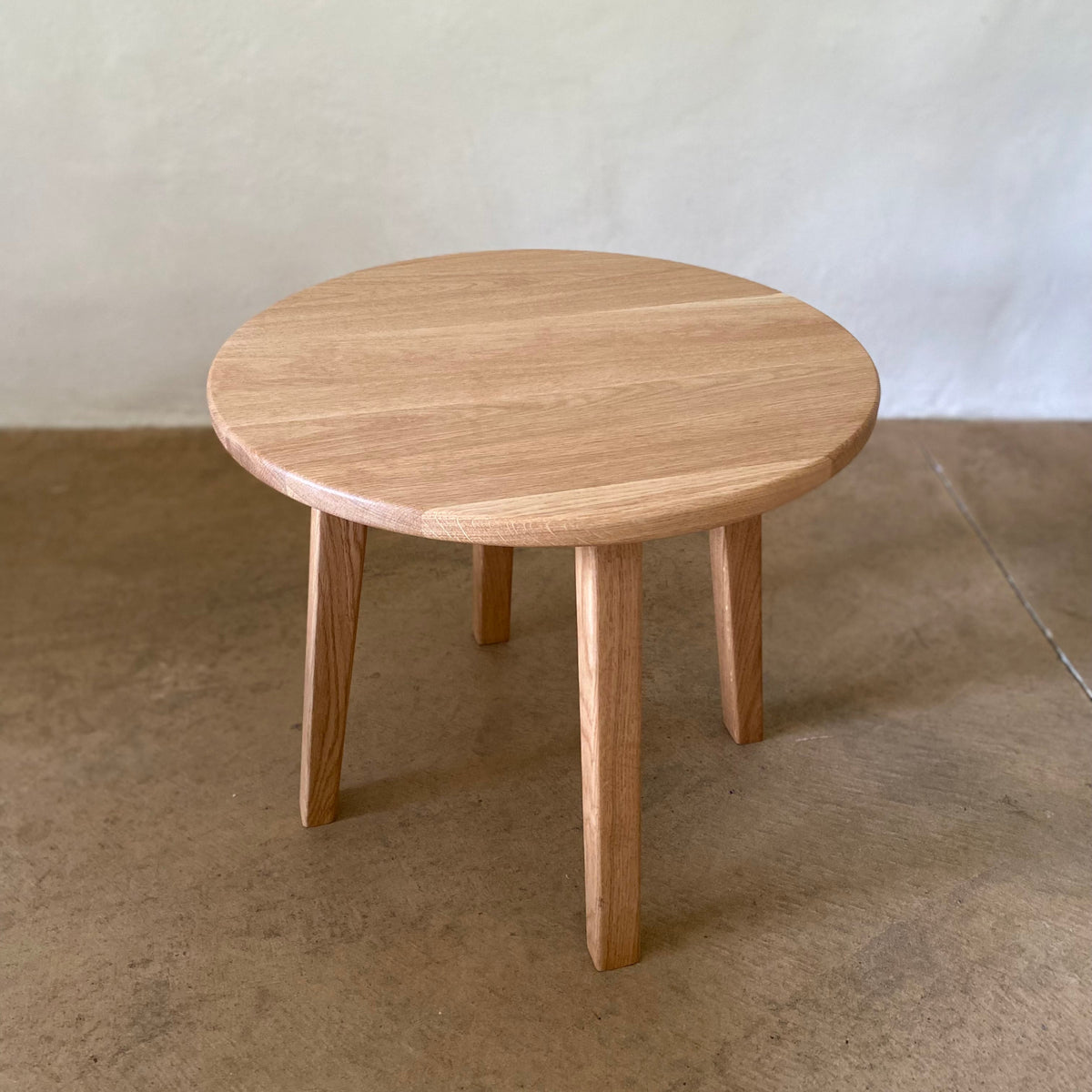 A round wooden side table with a light natural oak finish, featuring four tapered legs, set against a neutral concrete floor and textured wall.