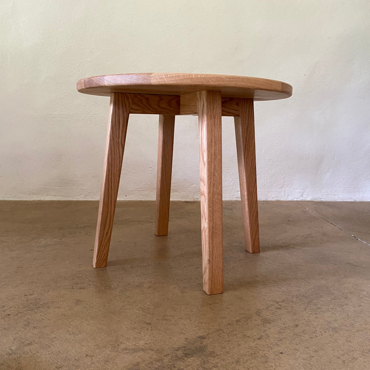 A small round wooden stool with four legs, shown from an angled overhead view against a split beige and concrete background.