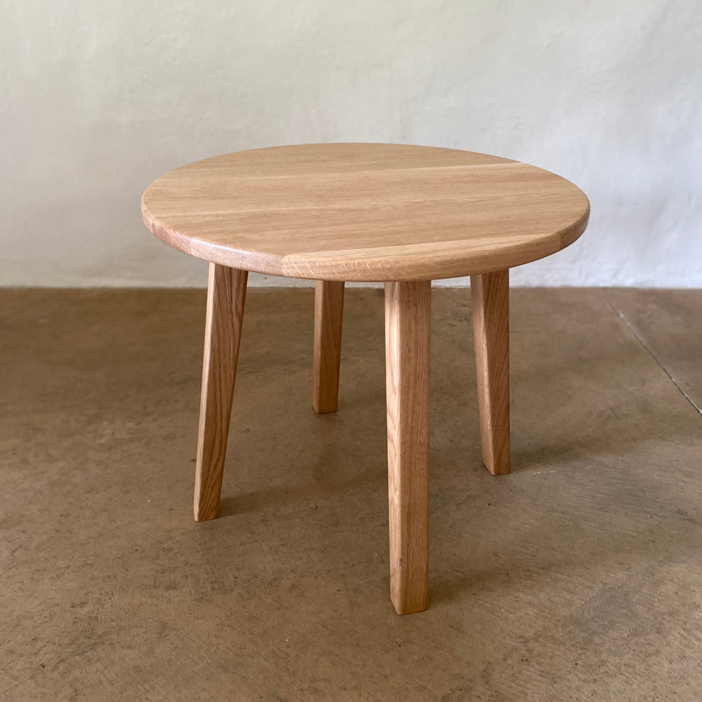A round wooden side table with four tapered legs, made of light natural wood, set against a textured off-white wall and concrete floor.