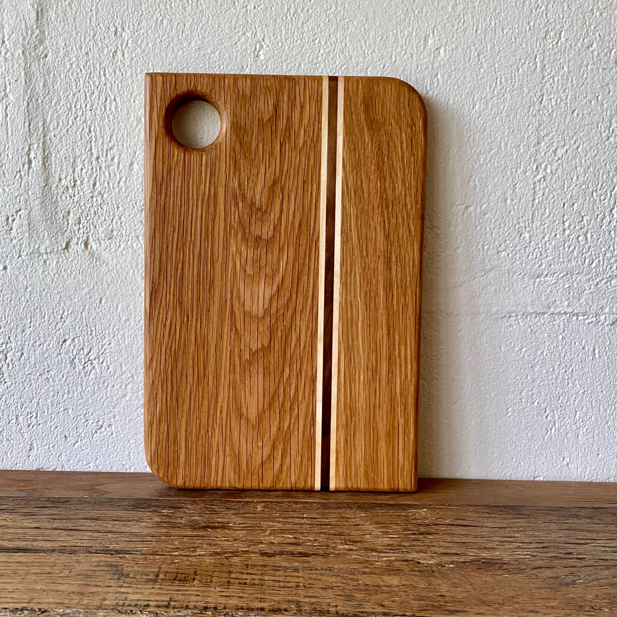 A rectangular white oak wooden cutting board with rounded corners, a circular hole near the top left, and a central inlay stripe, standing on a wooden surface against a textured white wall.