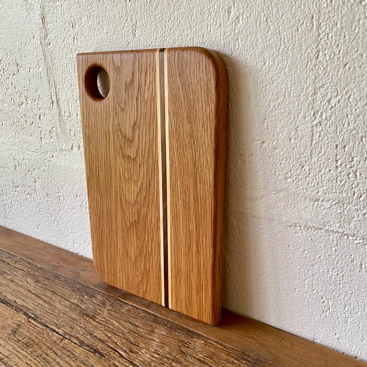 A rectangular white oak wooden cutting board with rounded corners, a circular hole near the top left, and a thin vertical inlay stripe, leaning against a textured off-white wall on a wooden surface.
