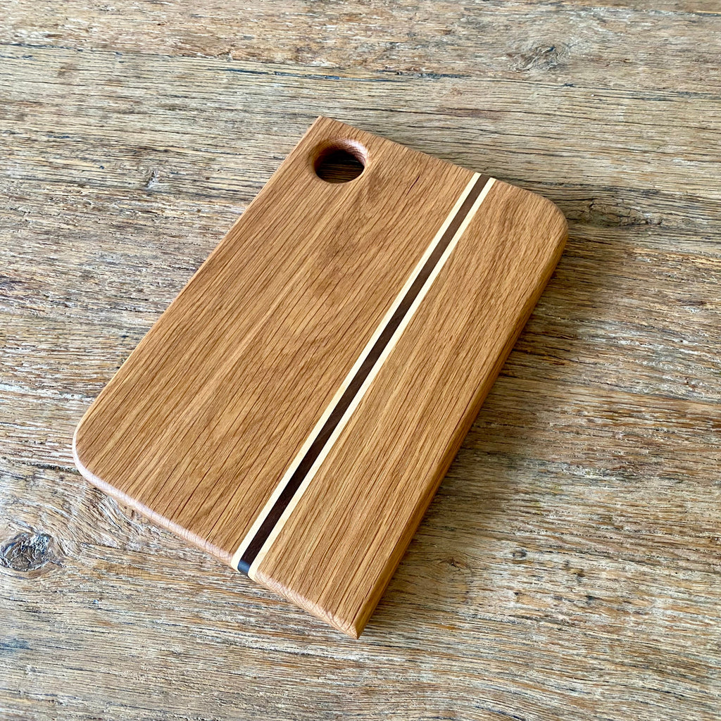 A rectangular wooden cutting board with rounded corners, featuring a natural oak finish, two parallel decorative inlay stripes, and a circular hanging hole in the top-left corner, shown on a rustic wooden tabletop.