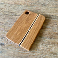A rectangular wooden cutting board with rounded corners, featuring a natural oak finish, two parallel decorative inlay stripes, and a circular hanging hole in the top-left corner, shown on a rustic wooden tabletop.