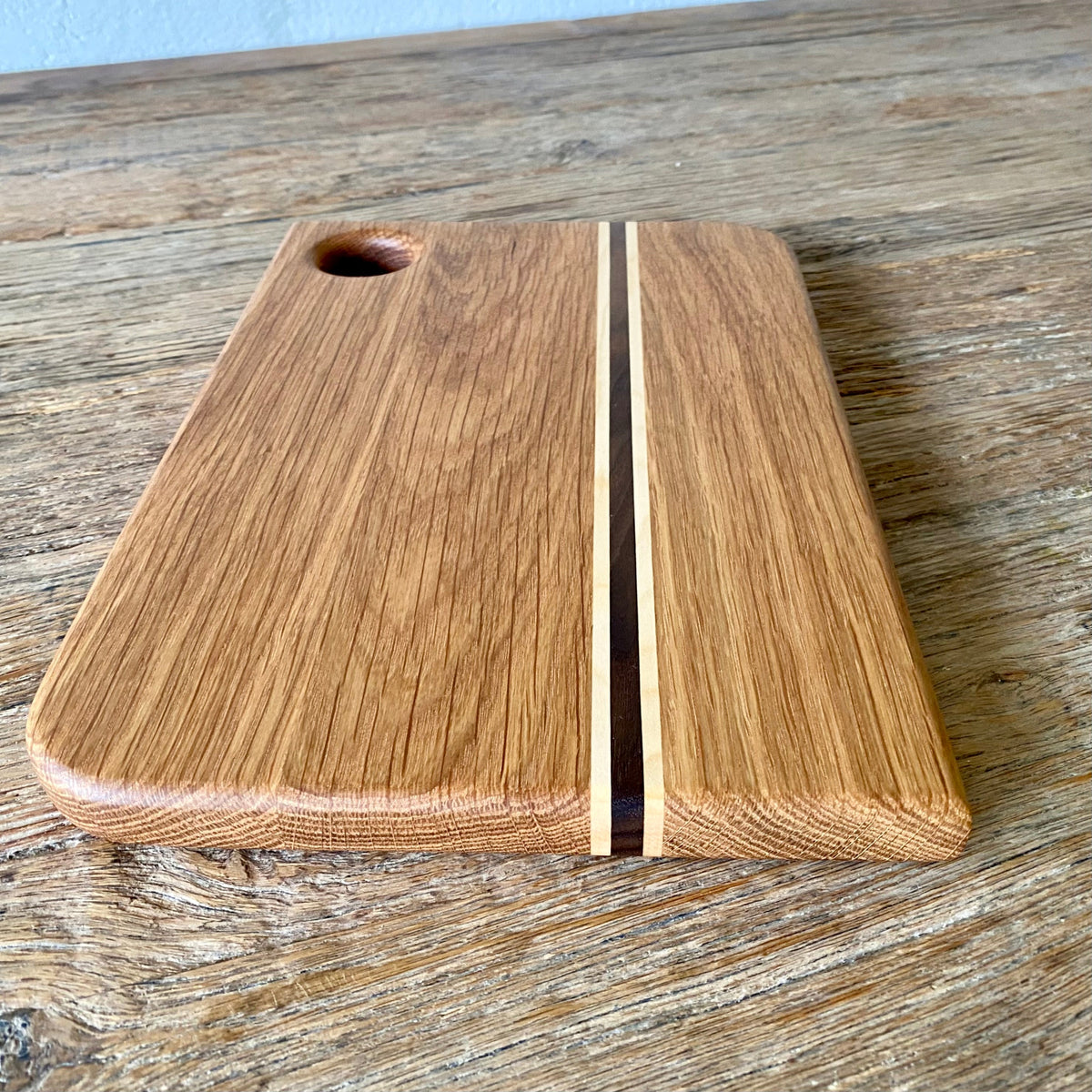 A rectangular white oak wooden cutting board with rounded corners, a central light and dark wood stripe, and a circular handle cutout, shown on a rustic wooden tabletop.