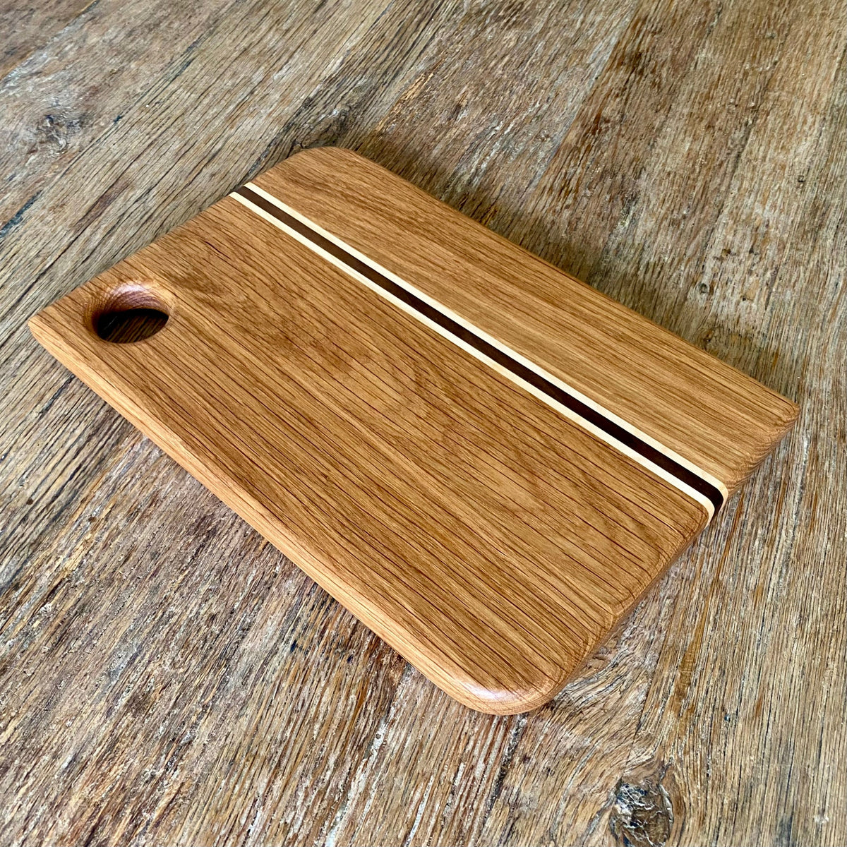 A rectangular white oak wooden cutting board with rounded corners, featuring a natural wood grain finish, a central decorative stripe with contrasting light and dark wood inlays, and a circular handle cutout on one end, shown on a rustic wooden tabletop.