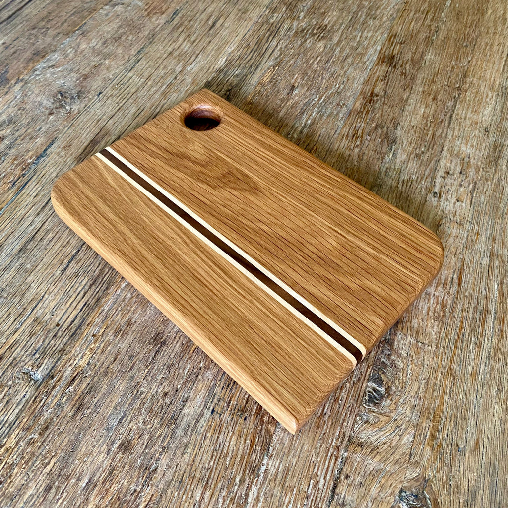 A rectangular wooden cutting board with rounded corners, featuring a natural oak finish, two decorative inlaid wood stripes, and a circular handle cutout, shown on a rustic wooden tabletop.